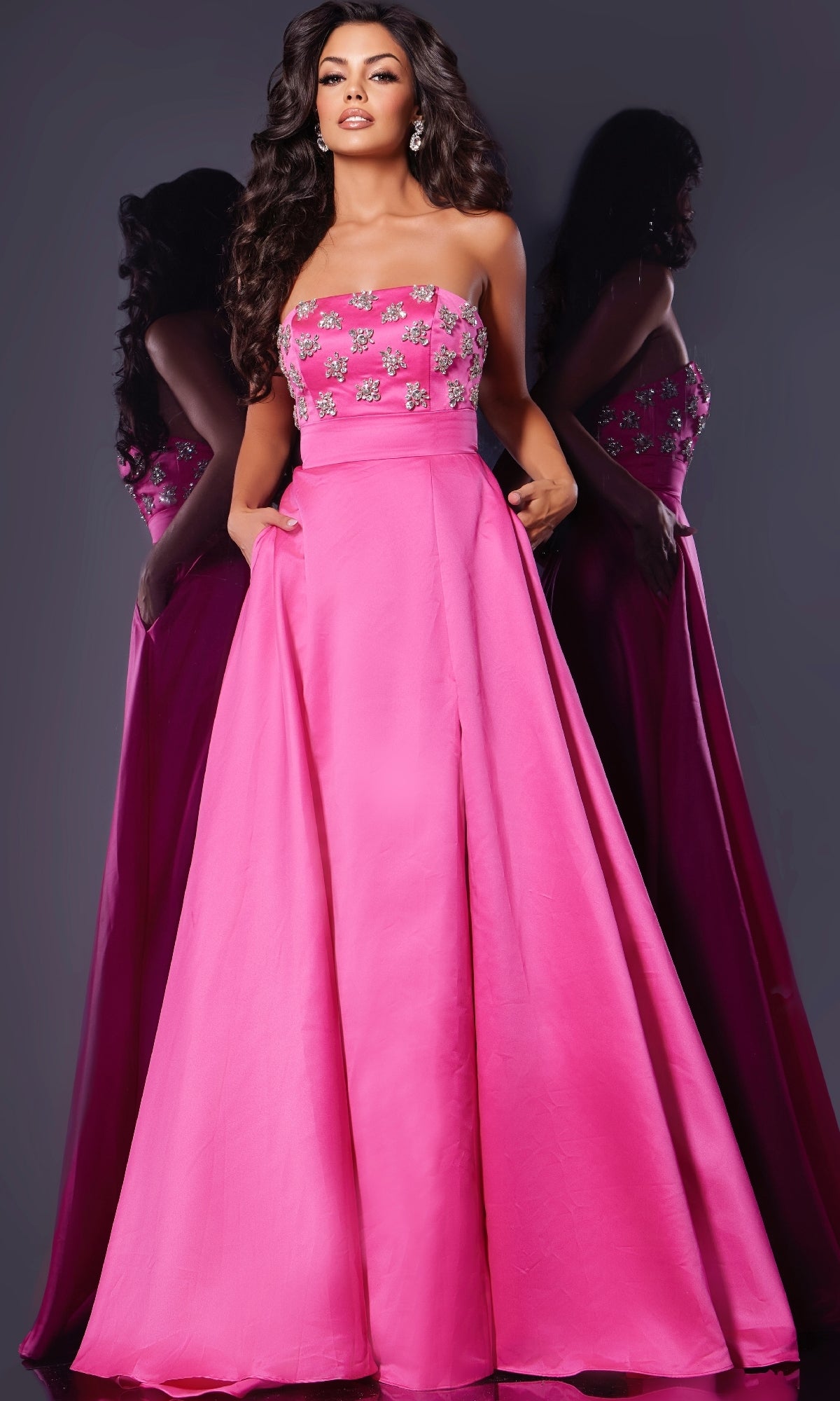 A woman stands against a reflective background wearing the Jovani 43822 long prom dress, a hot pink strapless gown with an A-line floor-length skirt and floral embellishments on the bodice.